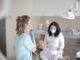 Femme allongée dans un fauteuil dentaire, une autre debout avec un masque, une lumière dentaire au-dessus